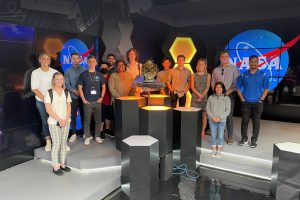 14 young people gather on a stage in front of black glass and NASA logos. In front hexagonal pedastles support a model of the James Webb Space Telescope, with it's golden honeycomb mirror. Hexagons and technical drawings permeate the design.