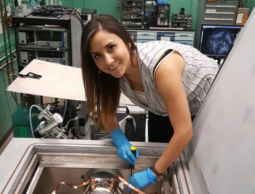 Margaret Dominguez, a woman with medium skin and long brown hair, smiles and leans over a table with copper wires and a round silver instrument.