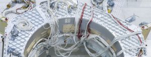 Seen from above, there is a view inside of a large, silvery metal structure with a hexagonal shape and a large cylindrical hole, covered in a diamond-patterned texture. Red and white wire bundles of cables drape across the top of the structure like strands of spaghetti.