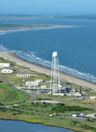 Aerial view of the coastal launch range of Wallops Flight Facility, showing a blue Atlantic Ocean on the right; white buildings along a tan coastline back up to a green, marshy landscape