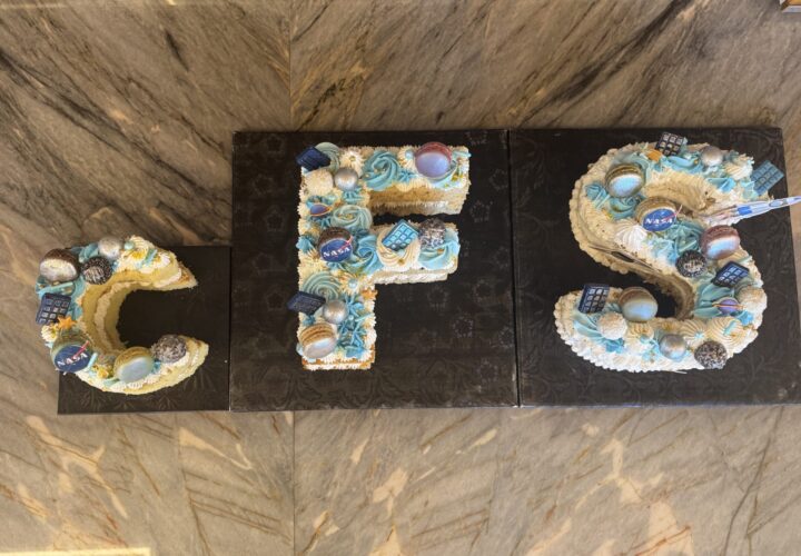 cFS logo-shaped celebration cake at the 2026 NASA cFS Symposium. Overhead view of a two-tiered cake shaped to spell 'cFS' in large letters on black bases. Each letter is decorated with golden frosting swirls, blue and orange fondant planets, NASA mission patches, and metallic spheres representing celestial bodies.